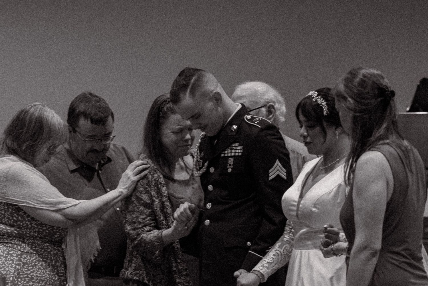 Military groom in dress uniform kisses bride during wedding prayer circle with family in black and white photo.