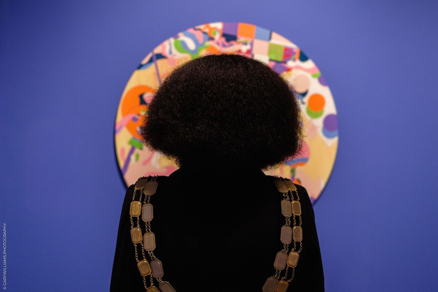 Person with curly bob hairstyle viewed from behind, standing before a colorful circular painting on a blue wall.
