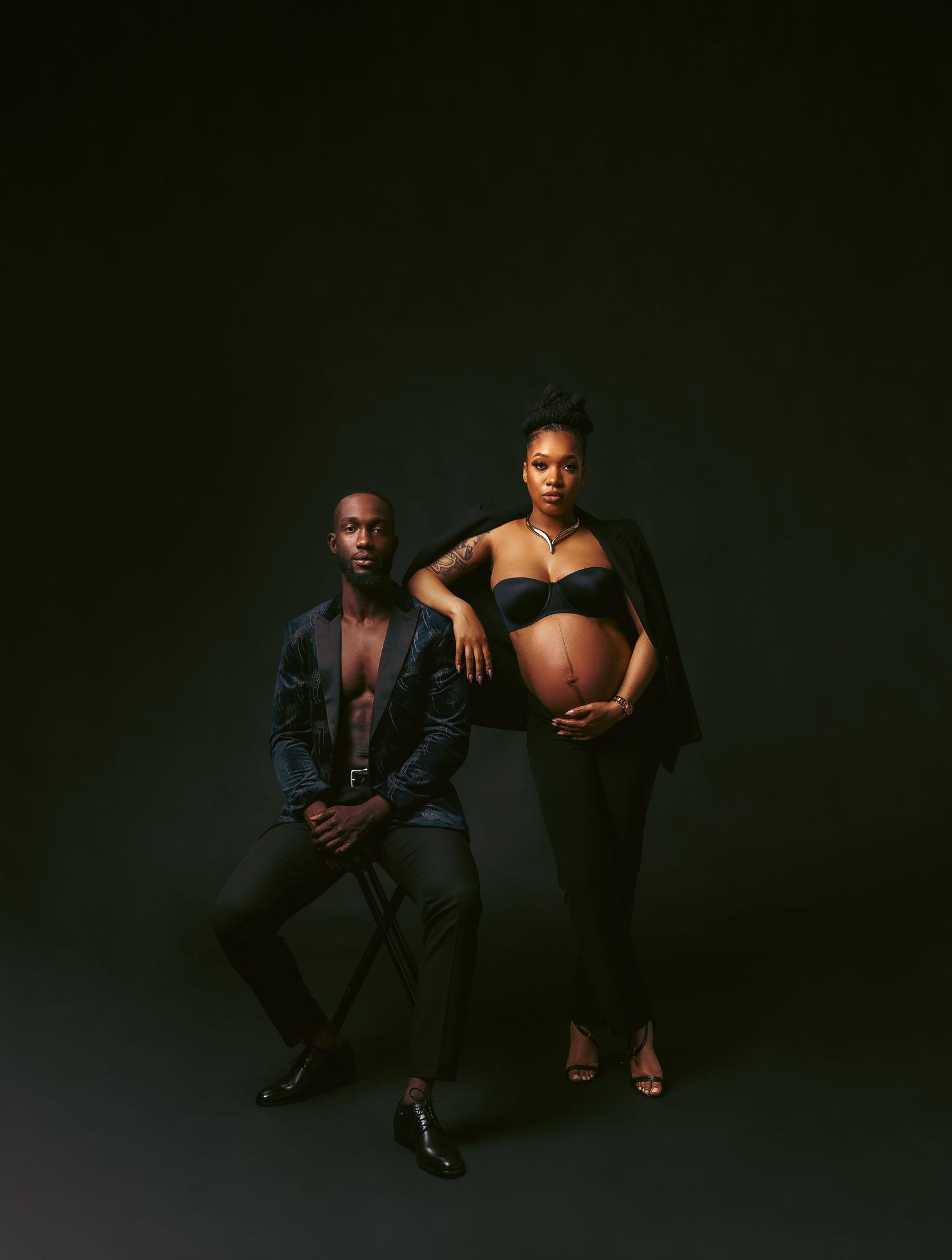 Couple poses dramatically in dark studio, man seated while pregnant woman stands in black outfit.