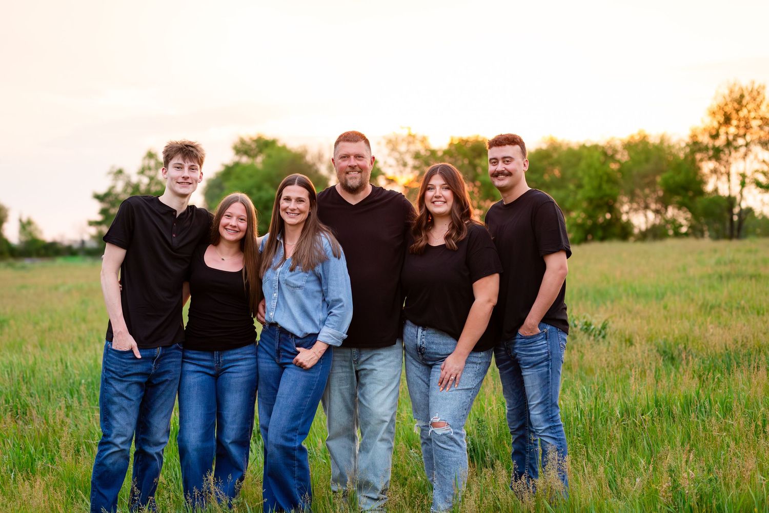 Family of six smiling together in a green field at sunset, wearing casual black tops and jeans.