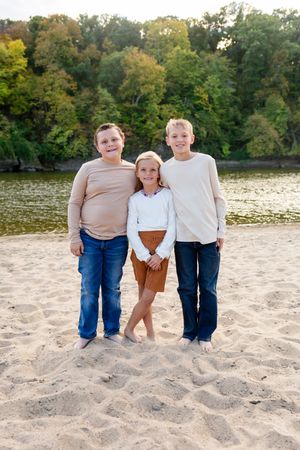 Three children smiling on a sandy beach with a river and green trees in the background on a sunny day.