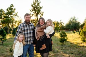 Family of four posing outdoors at a Christmas tree farm during golden hour, parents holding two young daughters.
