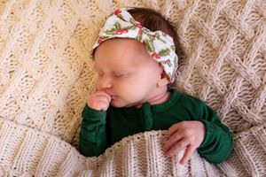 Sleeping newborn baby in green outfit and floral headband on cream knit blanket.