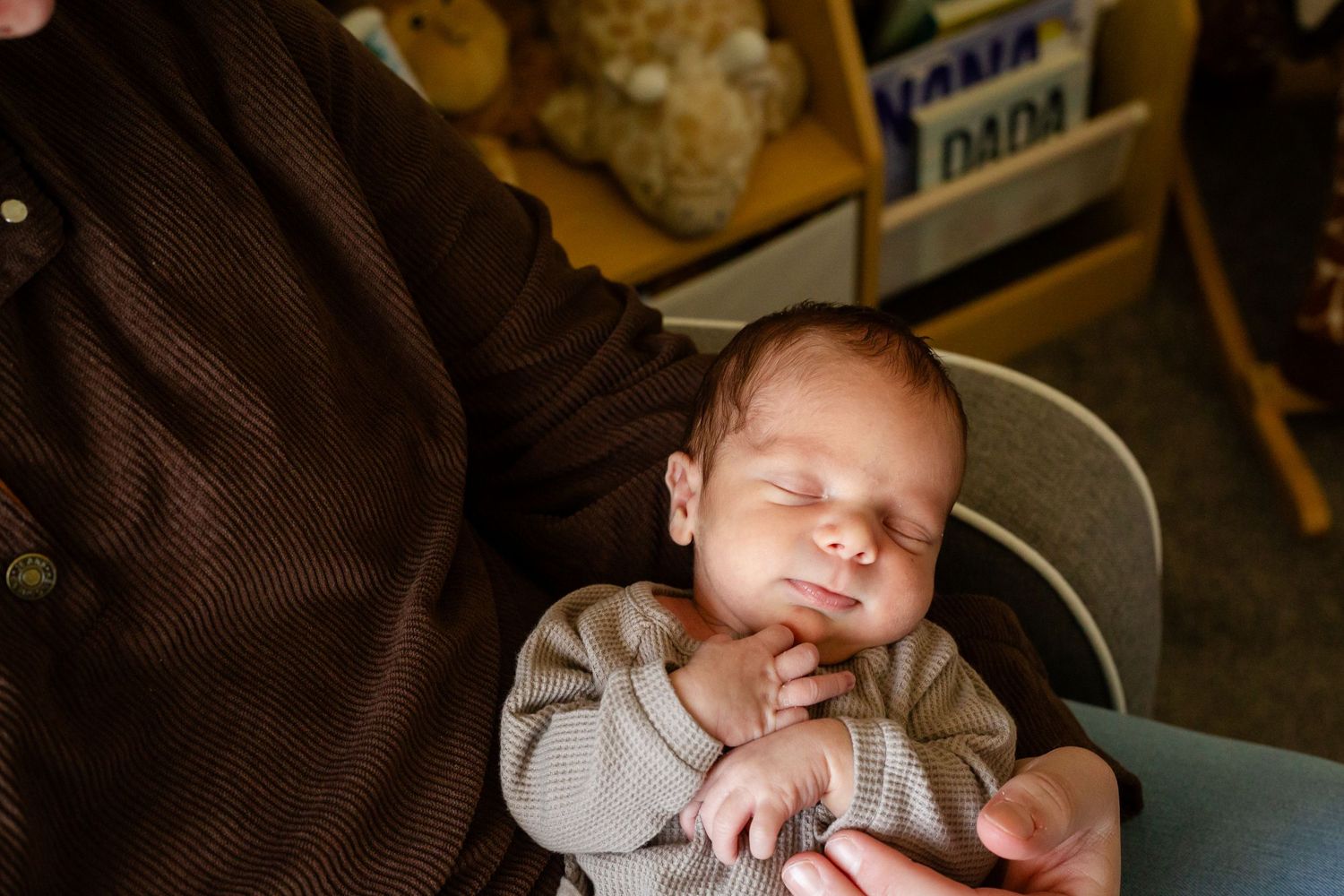 Newborn baby sleeping peacefully in adult hands, wearing a beige outfit, with teddy bears in the background.