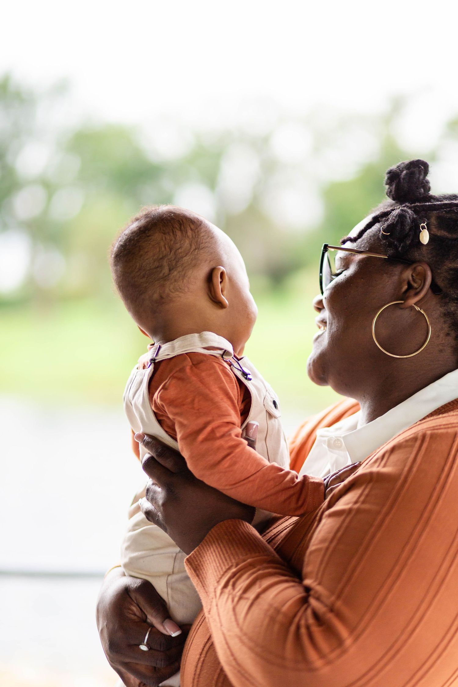 A smiling Black grandmother wearing glasses and hoop earrings holds a baby in an orange outfit near a bright window.