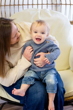 A joyful baby in a blue knit sweater and jeans sits on a mother's lap in a cozy white chair, laughing happily.