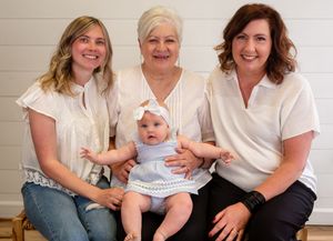 Three women and a baby girl in white tops pose together for a multi-generational family portrait photo.