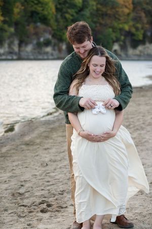Couple on beach, pregnant woman in white dress holding baby shoes on belly, man in green sweater embracing her from behind.