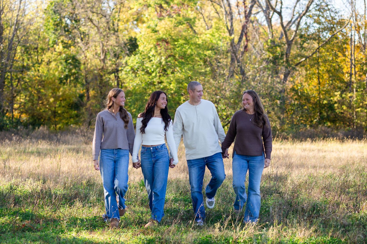 Family of four walking hand-in-hand through a sunlit meadow with autumn trees in the background.