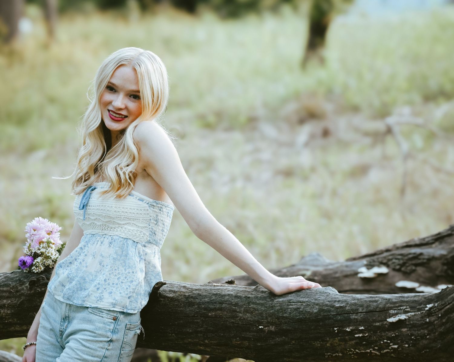 Young blonde woman in denim overalls holding wildflowers on a sunlit forest path.