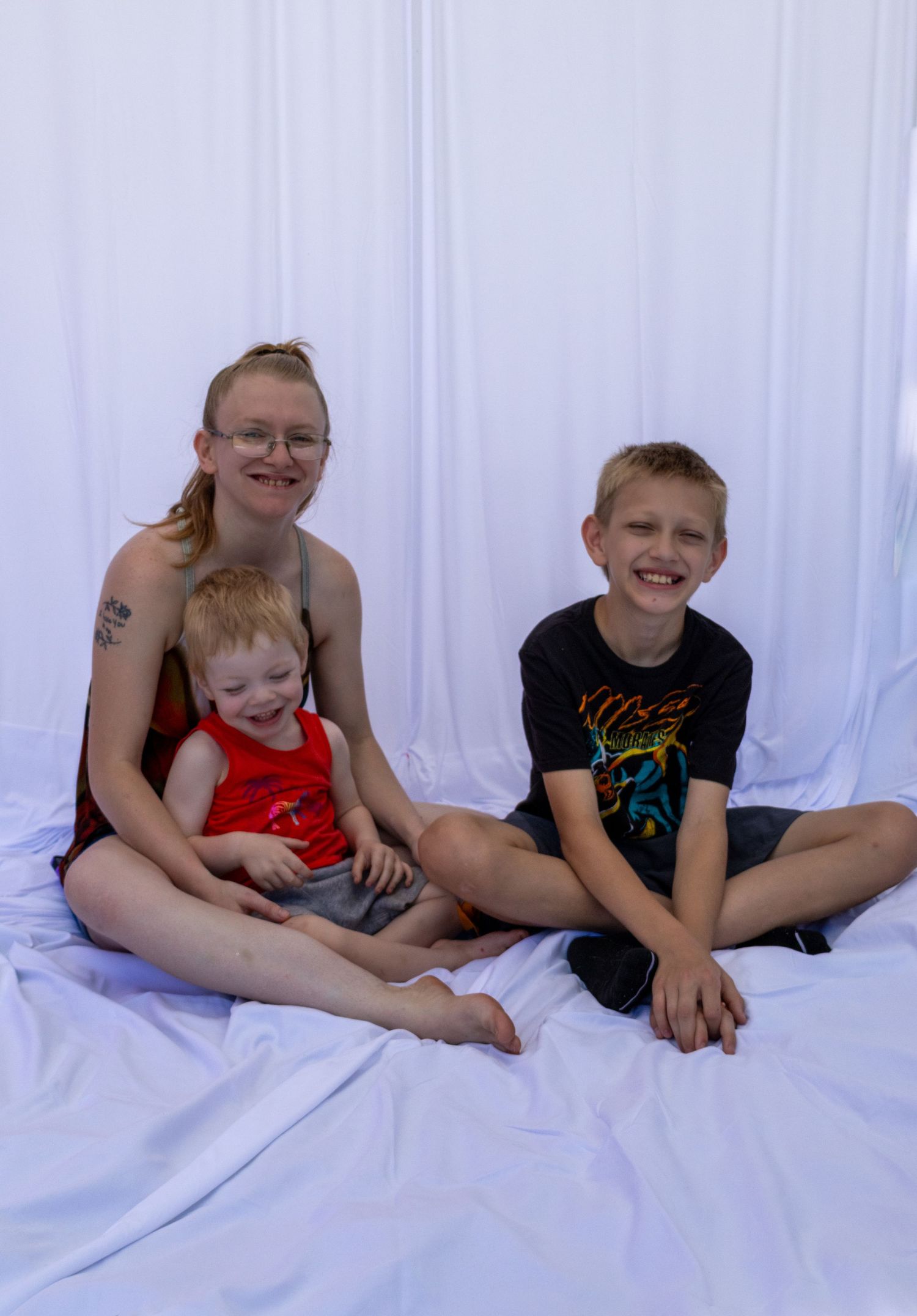 A woman with glasses sits cross-legged on white fabric with a toddler and a smiling boy in a black t-shirt.