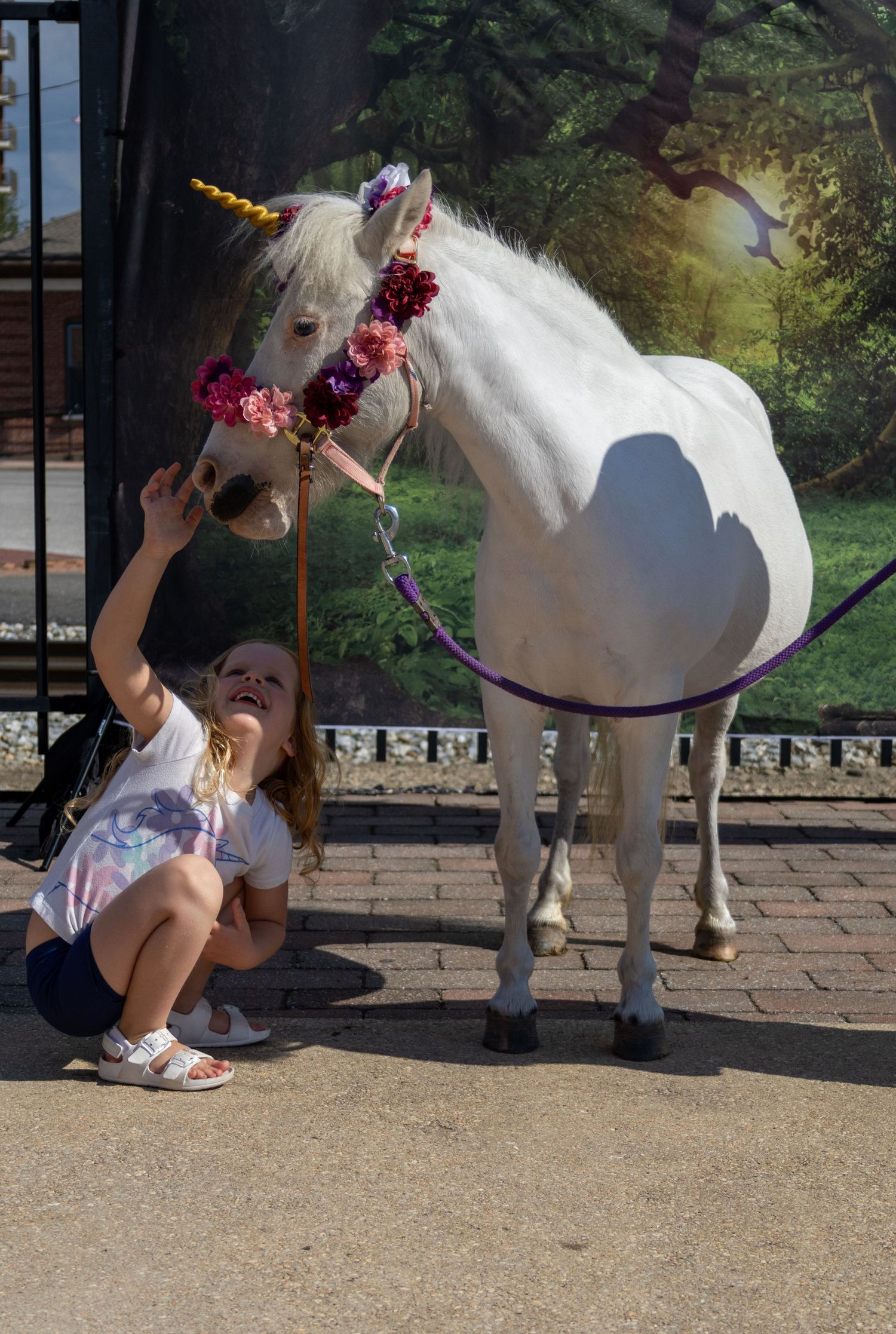 A young girl crouches beside a white horse decorated with a floral crown and golden unicorn horn outdoors.