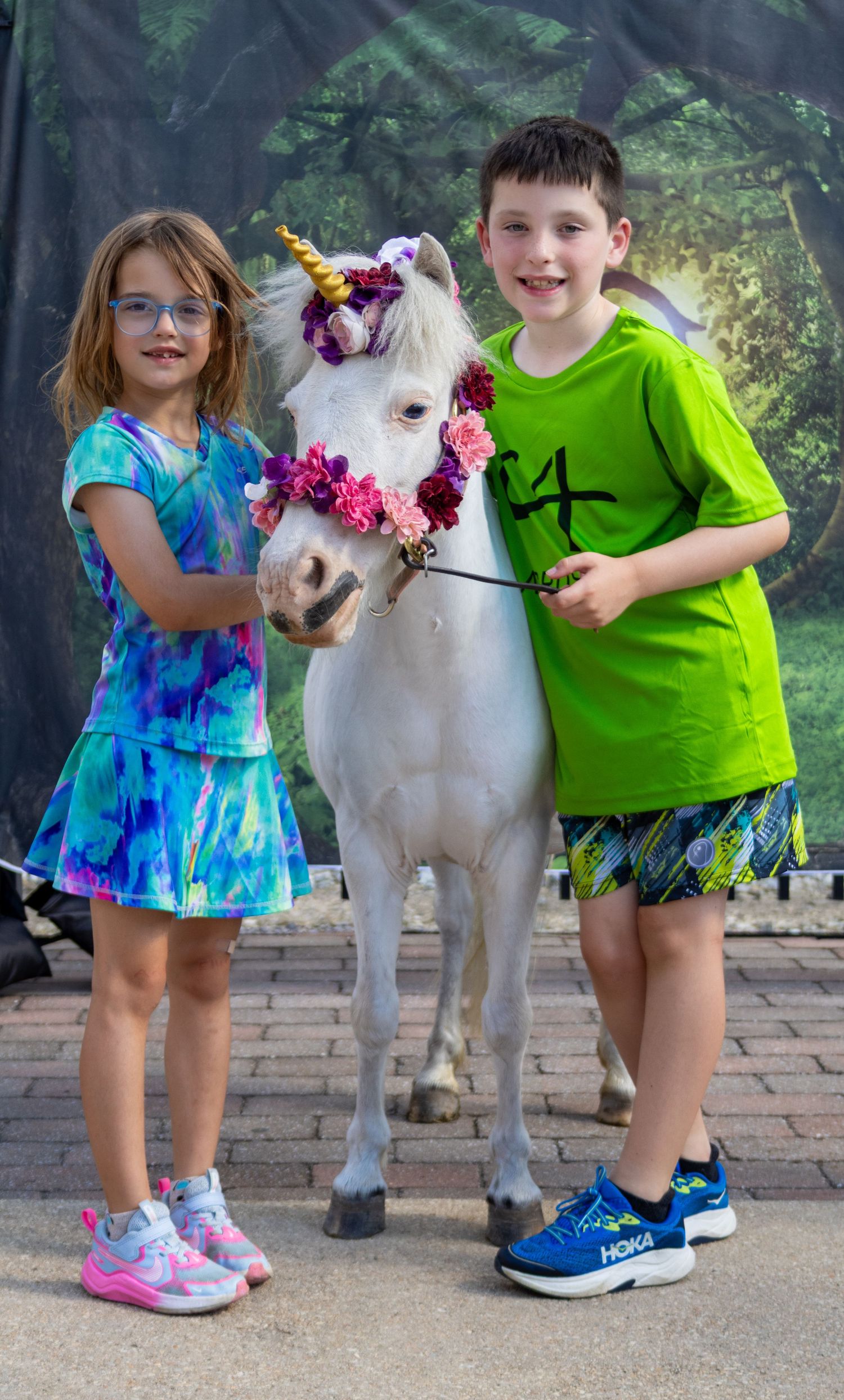 Two children pose with a white miniature horse decorated as a unicorn with floral crown at an outdoor event.