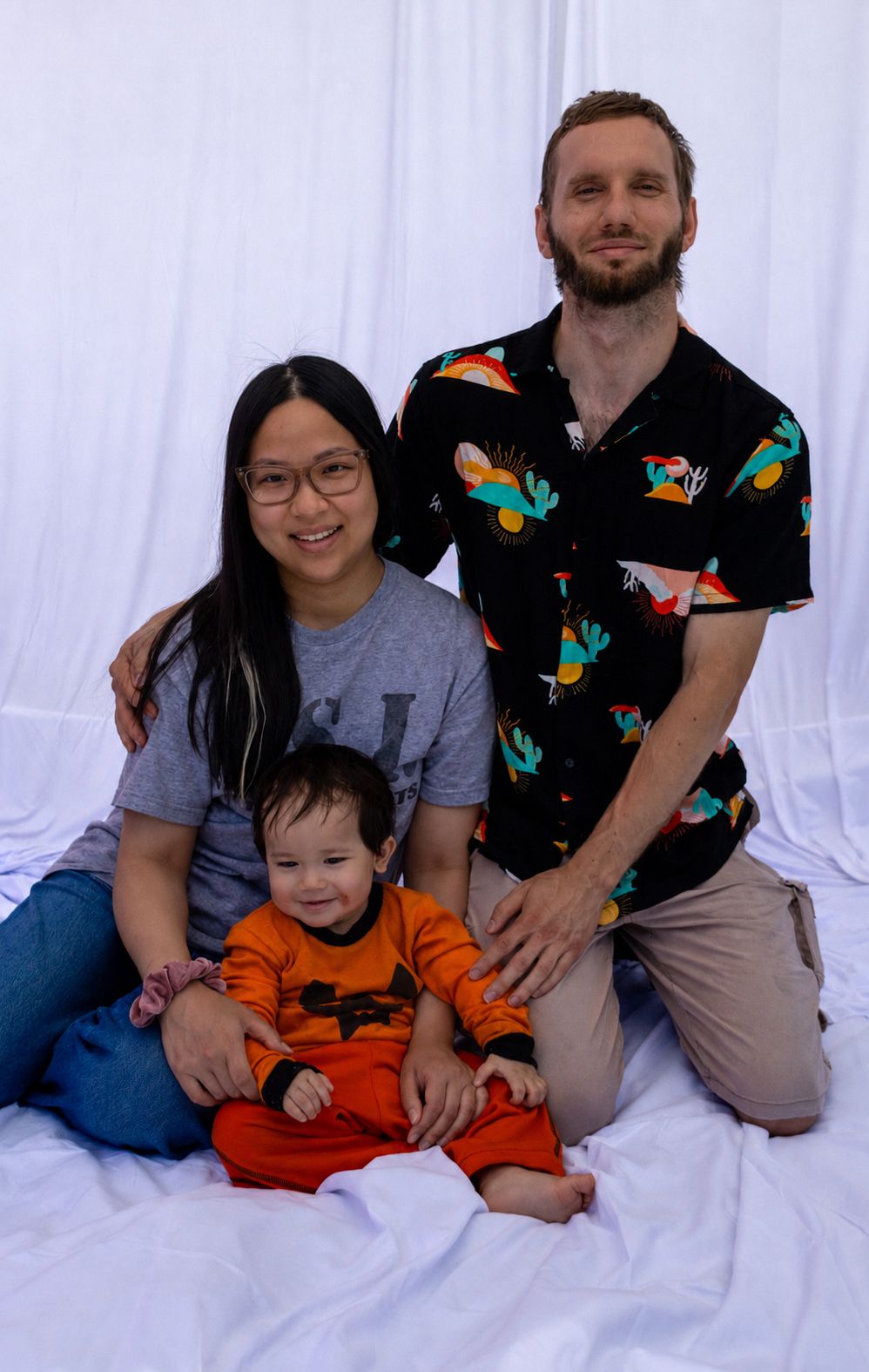 A young family poses for a portrait, with a woman, man, and baby in an orange outfit on a white backdrop.