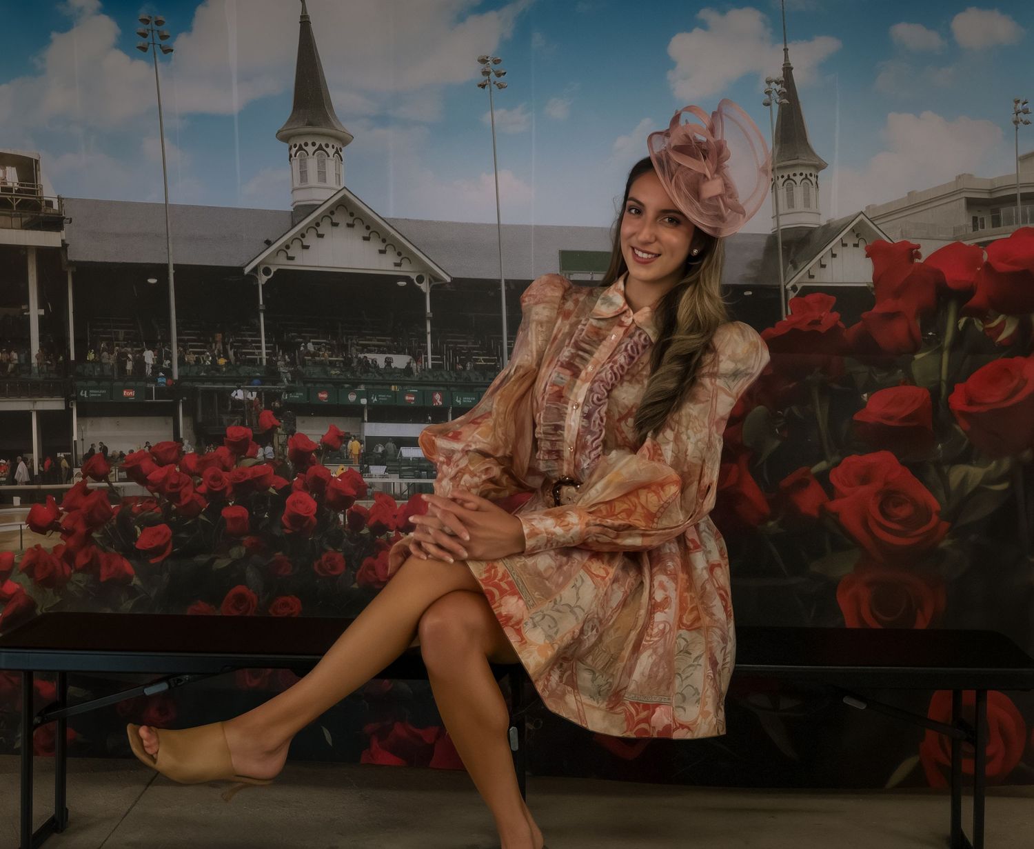 Woman in floral dress and pink fascinator poses at Kentucky Derby backdrop with twin spires and roses.