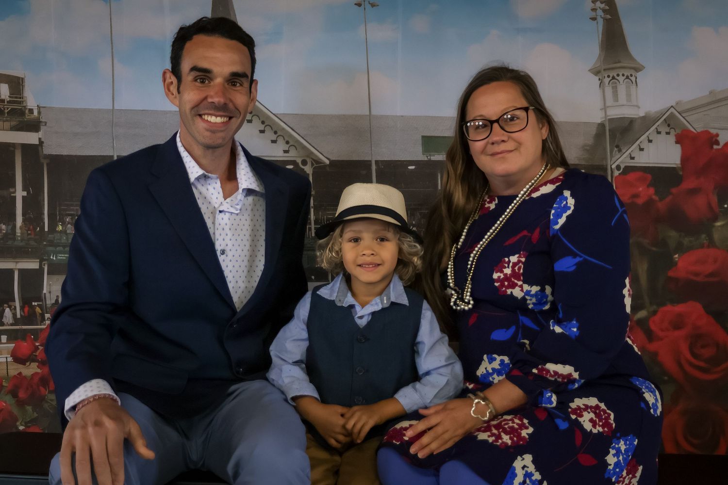 Family of three poses at Churchill Downs, child wearing a hat flanked by parents in formal attire.
