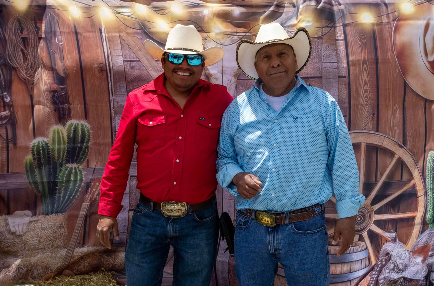 Two men in cowboy hats and western shirts pose in front of a rustic western-themed backdrop with cacti.