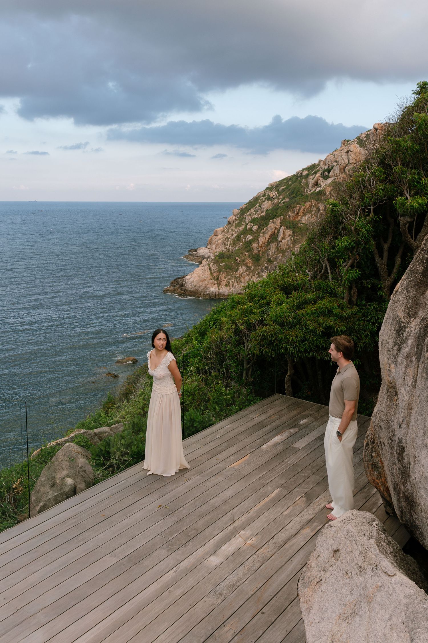 Couple standing on a wooden deck overlooking a rocky coastline with lush greenery and calm ocean views at dusk.