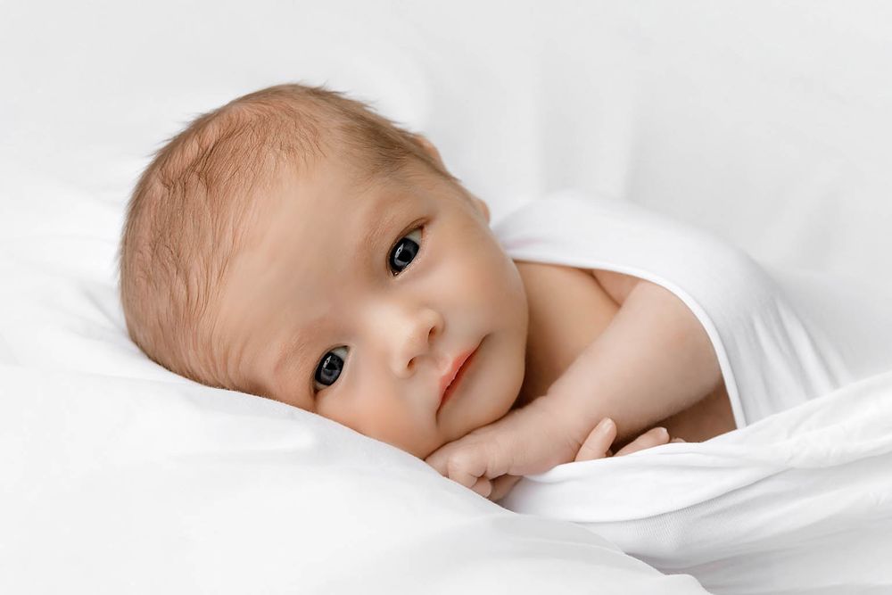 Newborn baby wrapped in white swaddle lying on white blanket, looking at camera with bright eyes.