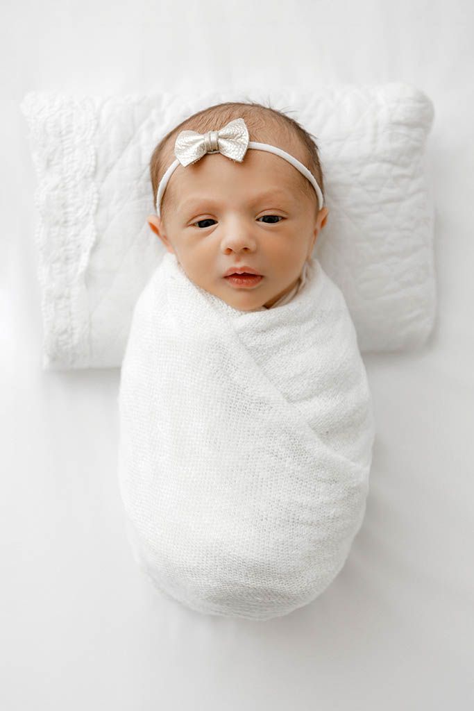 Newborn baby girl swaddled in white wrap with bow headband, lying on white pillow.
