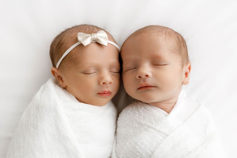 Two sleeping newborn twins swaddled in white blankets, one wearing a bow headband by Temecula newborn twins photographer.