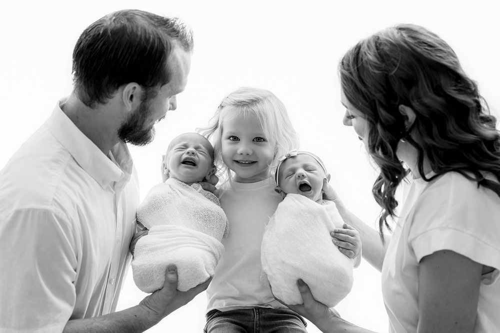 Black and white family photo of parents holding newborn twins with smiling toddler by Temecula newborn twins photographer.