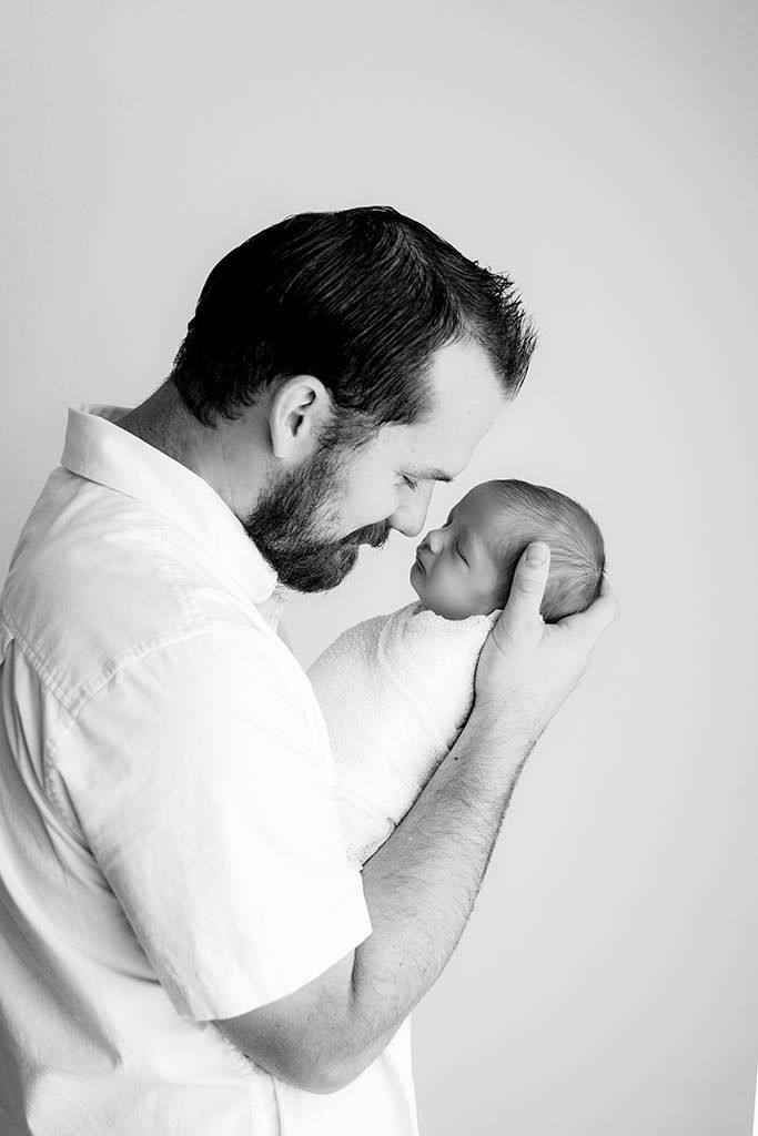 Black and white photo of a bearded father tenderly touching noses with his newborn baby.