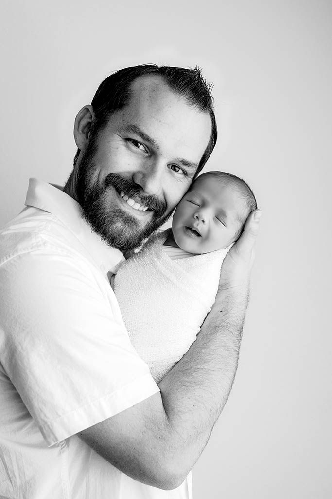 Black and white portrait of a smiling bearded man holding a sleeping newborn baby wrapped in white.