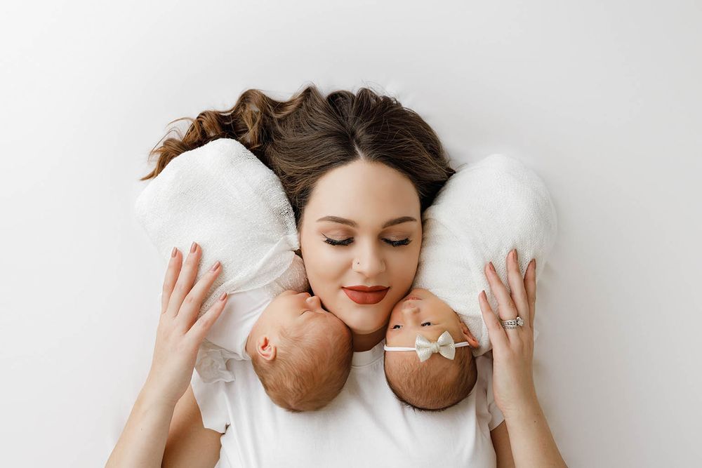 Mother lying down with eyes closed, tenderly holding two newborn babies on her chest against white background.