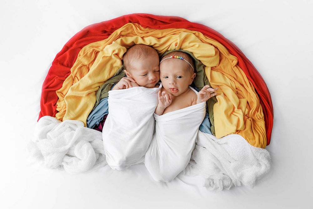Two swaddled newborn twins lying together inside a colorful rainbow-shaped fabric prop by Temecula newborn twins photographer.