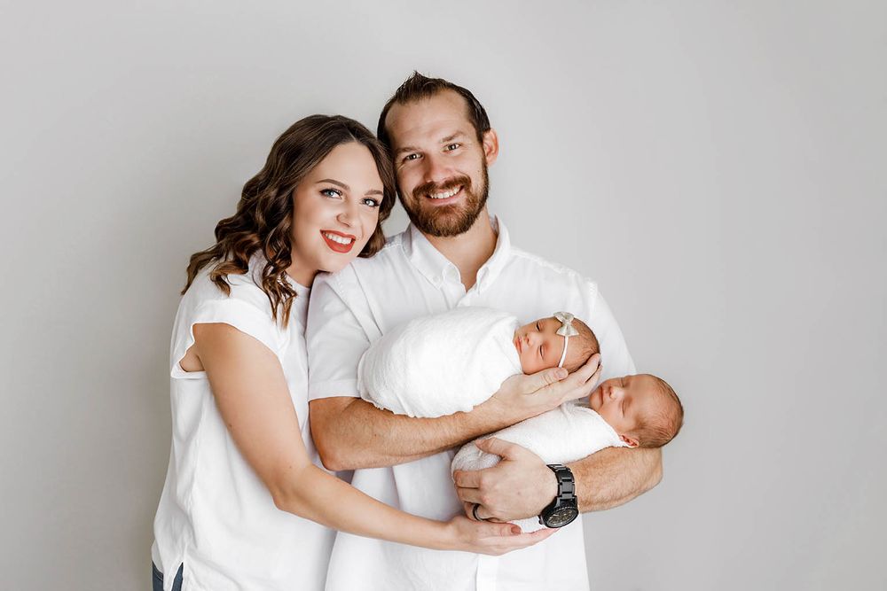 Happy couple in white outfits holding their newborn babies in a studio portrait.