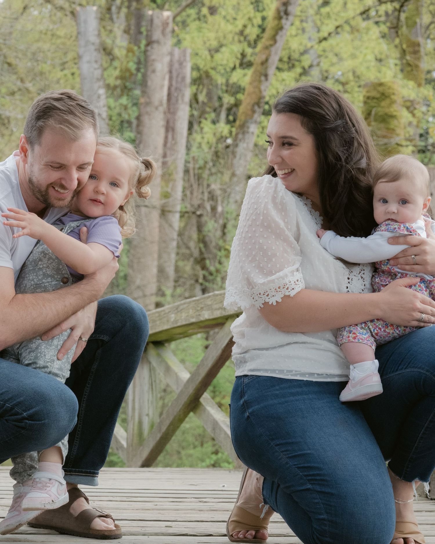A happy family of four posing on a bridge at Jericho Beach in Vancouver. The father holds a toddler girl, and the mother holds a baby.