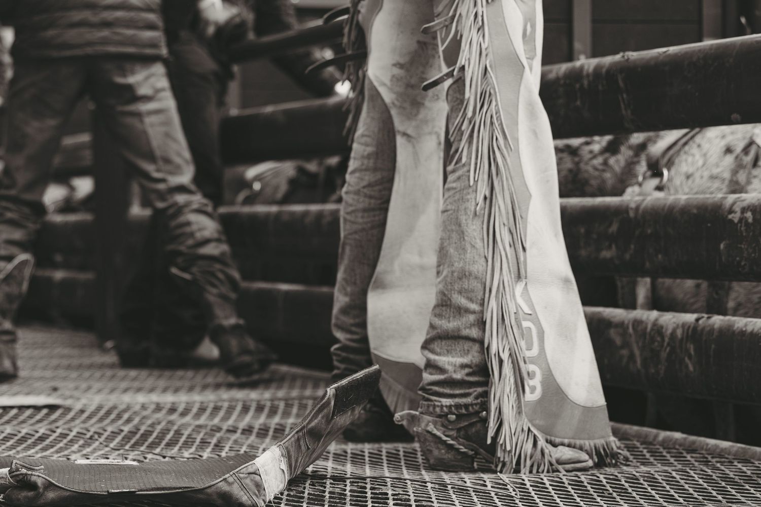 Black and white close-up of cowboy boots and fringed chaps at a rodeo event.