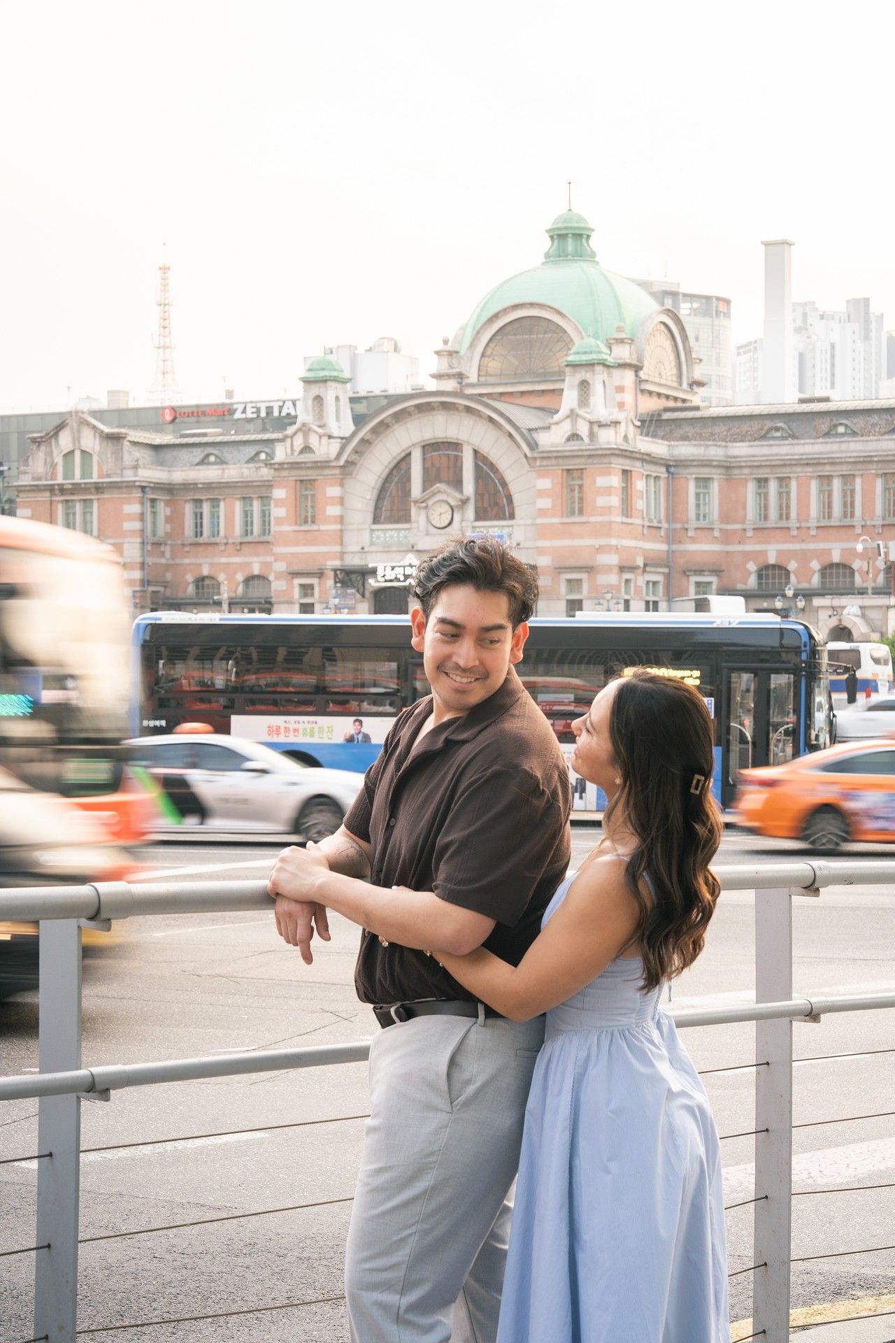 Couple posing romantically in front of Seoul Station's historic red brick building with city traffic in background.