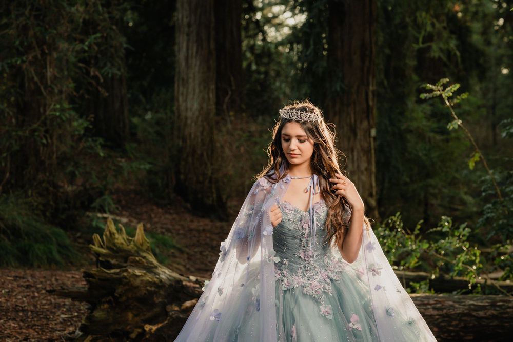 Young woman in a light blue ball gown with floral cape and crown poses in an enchanted forest setting.