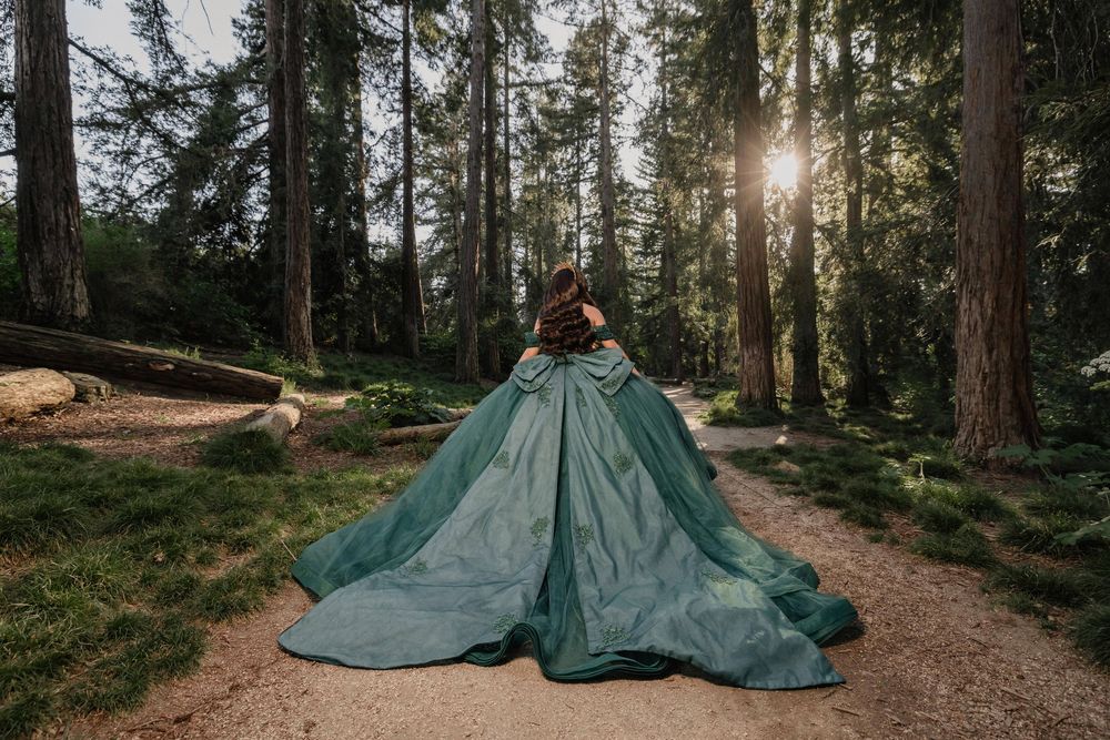 Woman in elegant teal ballgown walks forest path with sunlight filtering through tall trees.