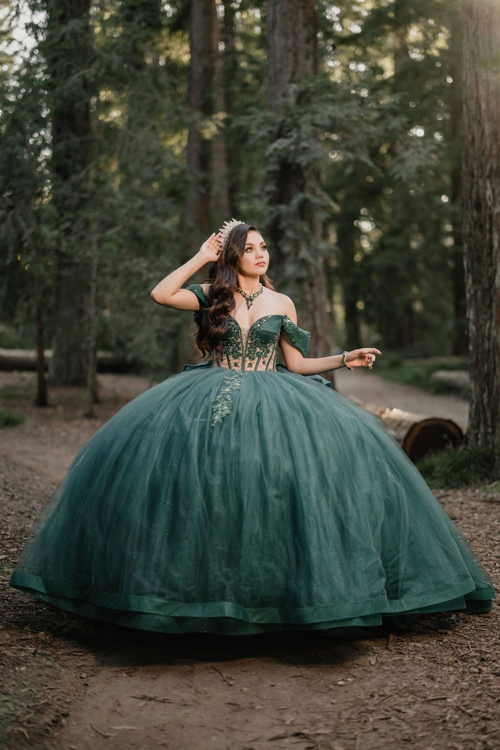 Young woman in an elegant emerald green ball gown with gold embroidery poses gracefully in a forest setting.