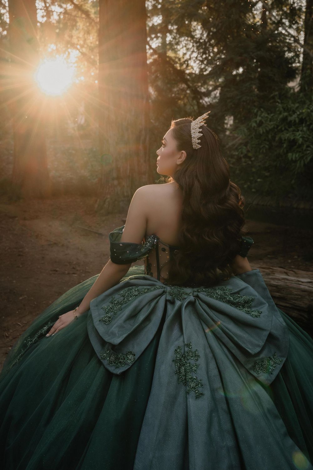 Woman in emerald green ball gown with crown poses in sunlit forest at golden hour.