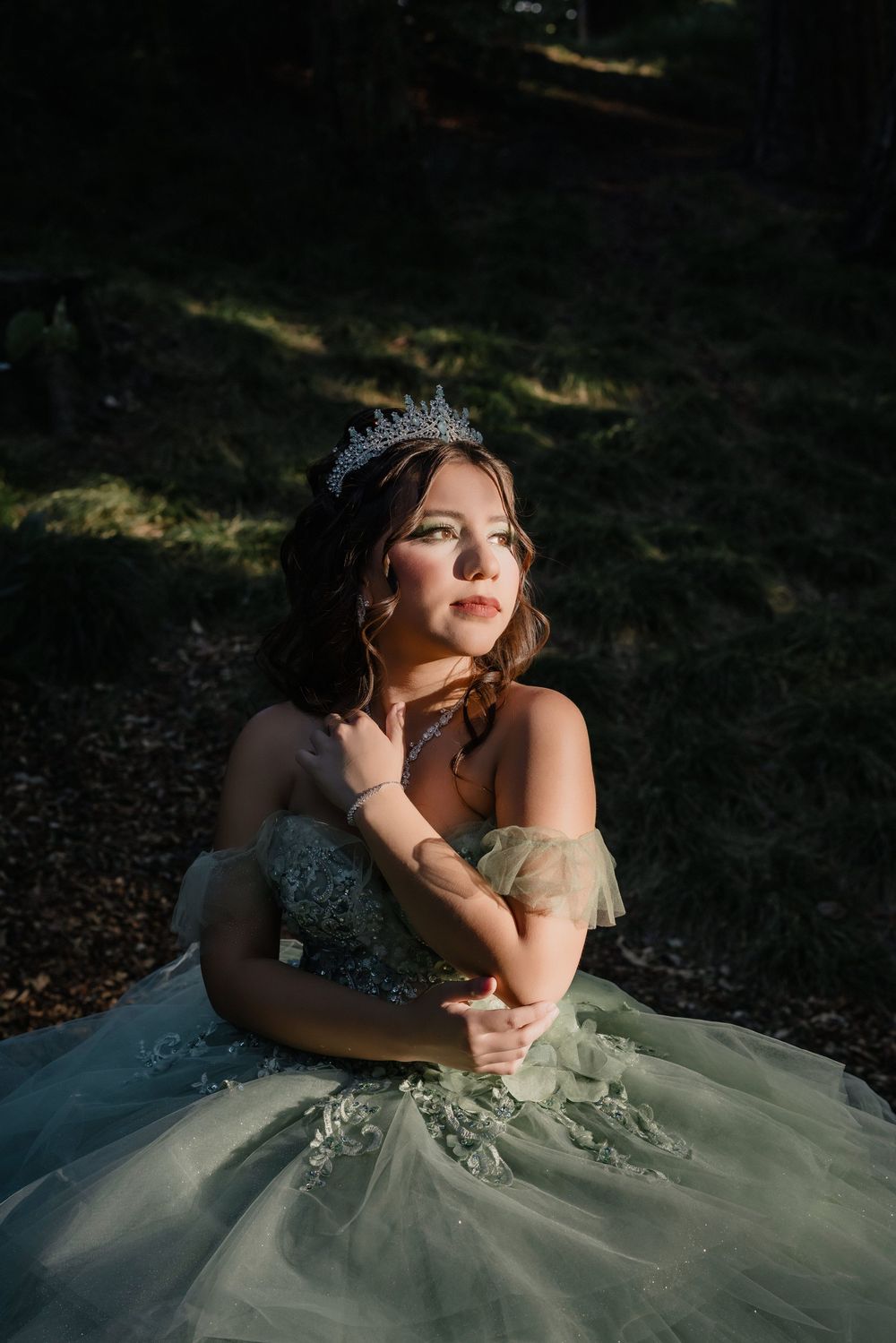 Young woman in sage green ball gown and silver tiara poses gracefully outdoors in dappled sunlight.