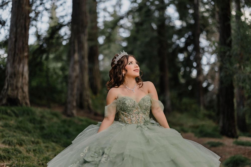 Young woman in sage green ball gown and tiara poses gracefully in a forest setting for quinceañera photos.