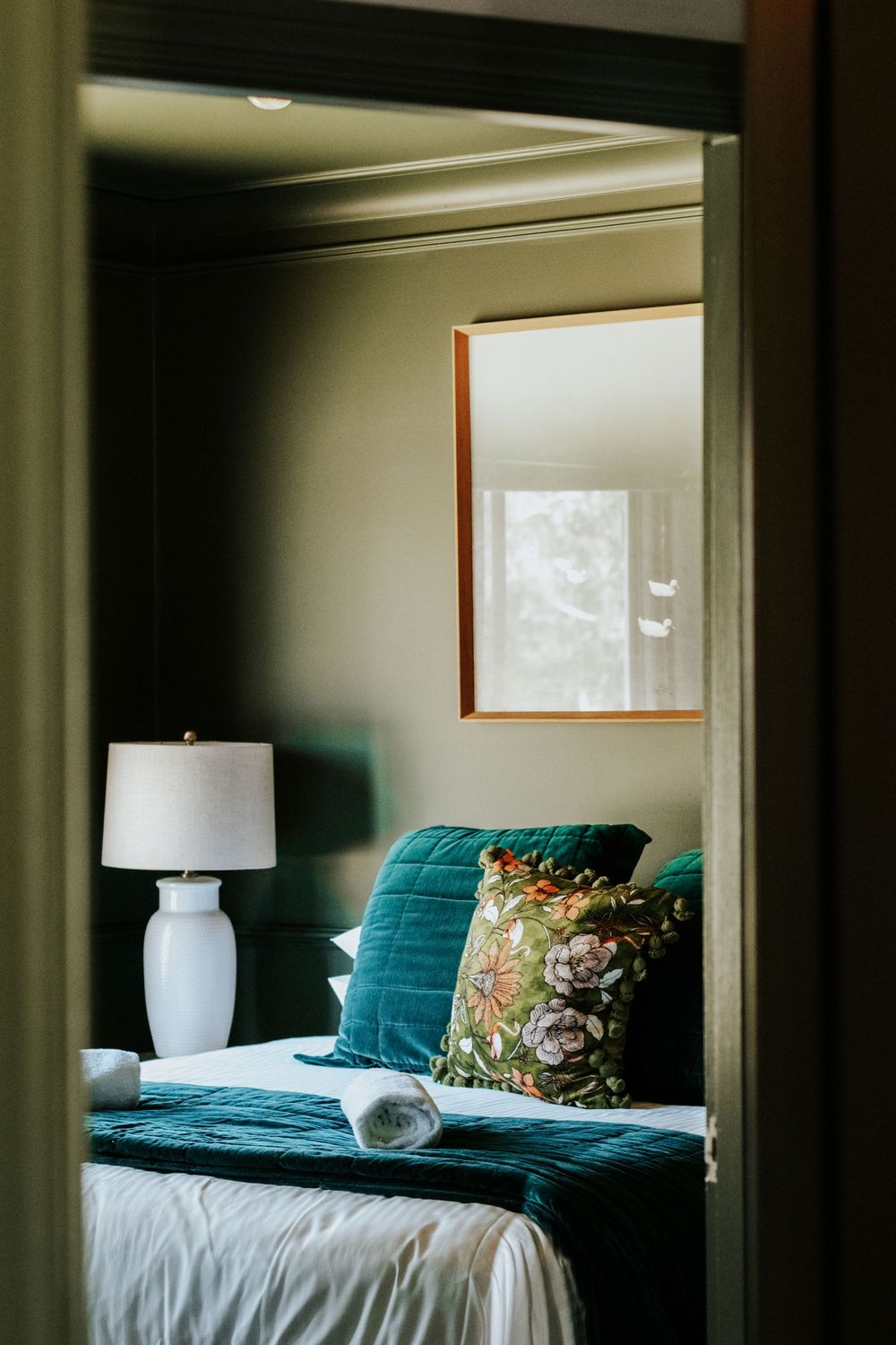 Cozy bedroom with teal velvet bedding, floral pillow, white lamp, and framed window viewed through doorway.