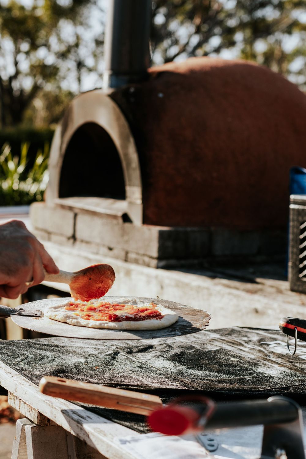 Person spreading tomato sauce on pizza dough on a wooden peel near a traditional outdoor wood-fired clay oven.