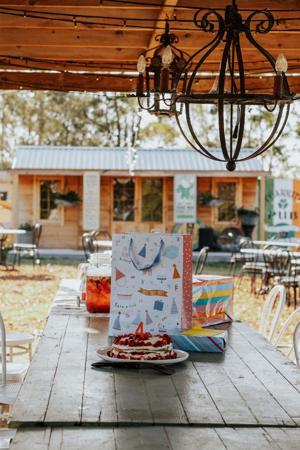 Outdoor birthday party table with cake, gift bags, and drinks under a chandelier, with a rustic wooden cabin in the background.