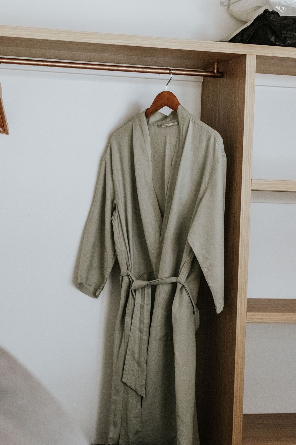 Sage green linen robe hanging on a wooden hanger inside a minimalist open wardrobe closet.