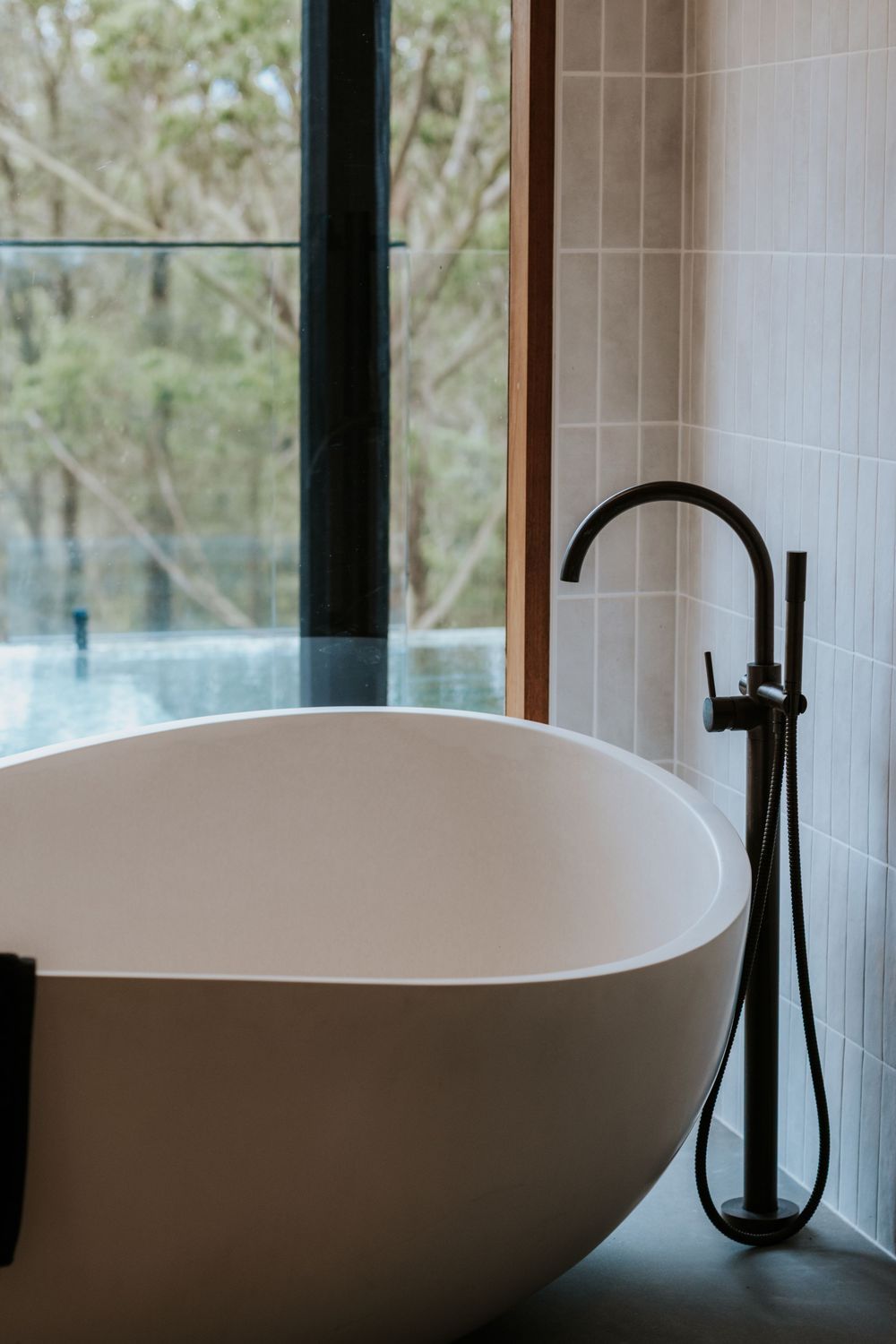Modern freestanding white oval bathtub with matte black floor-mounted faucet beside large window overlooking lush greenery.