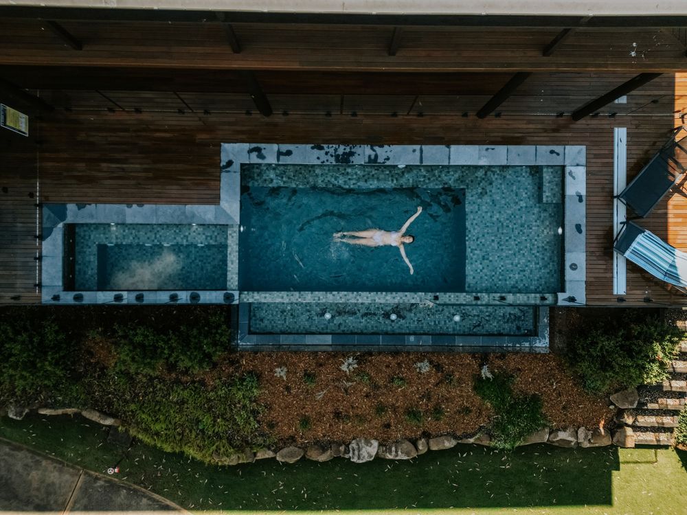 Aerial view of a person floating in a rectangular outdoor pool surrounded by wooden decking and lush greenery.