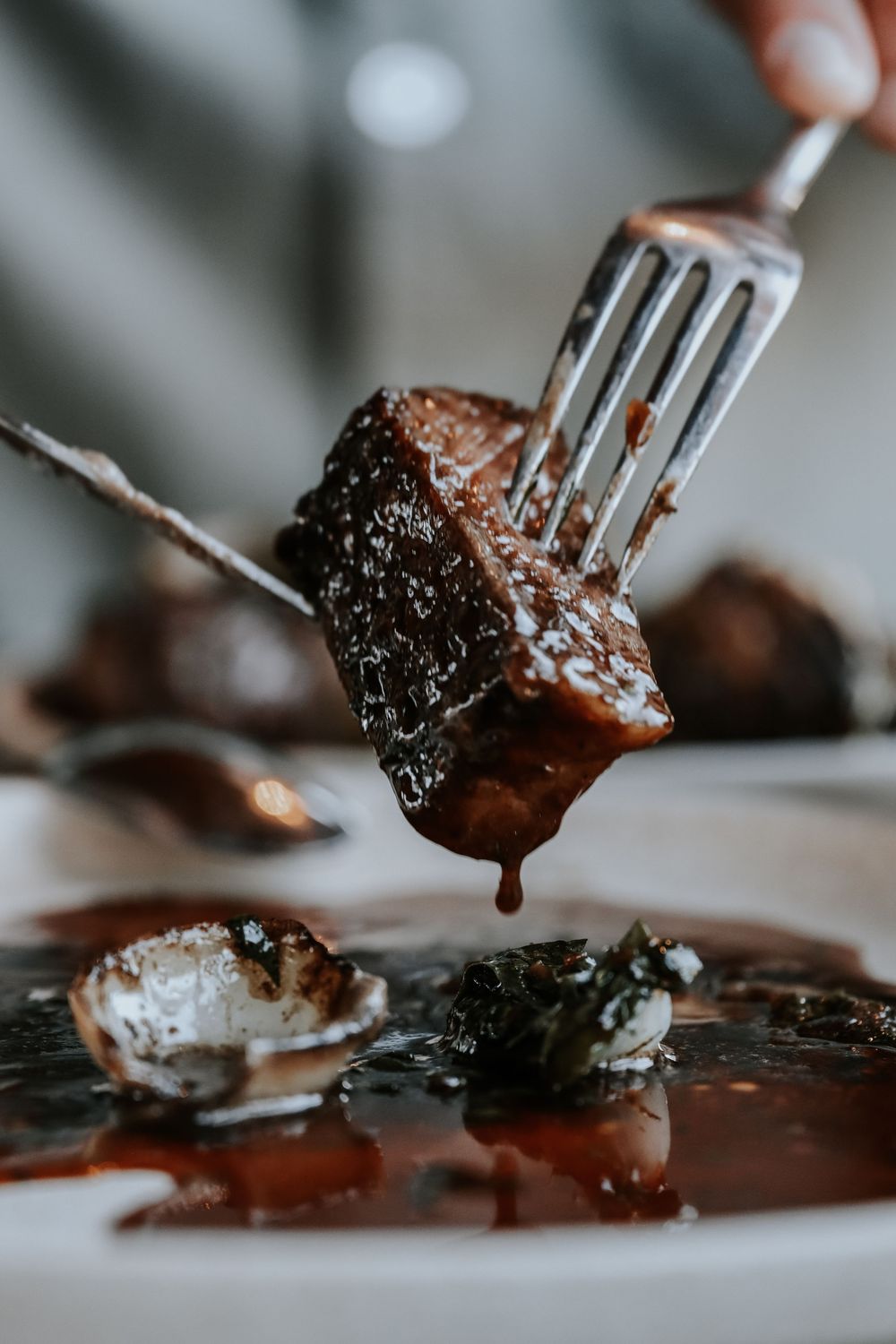 A fork lifting a glazed, caramelized meat piece dripping rich brown sauce over a white plate.