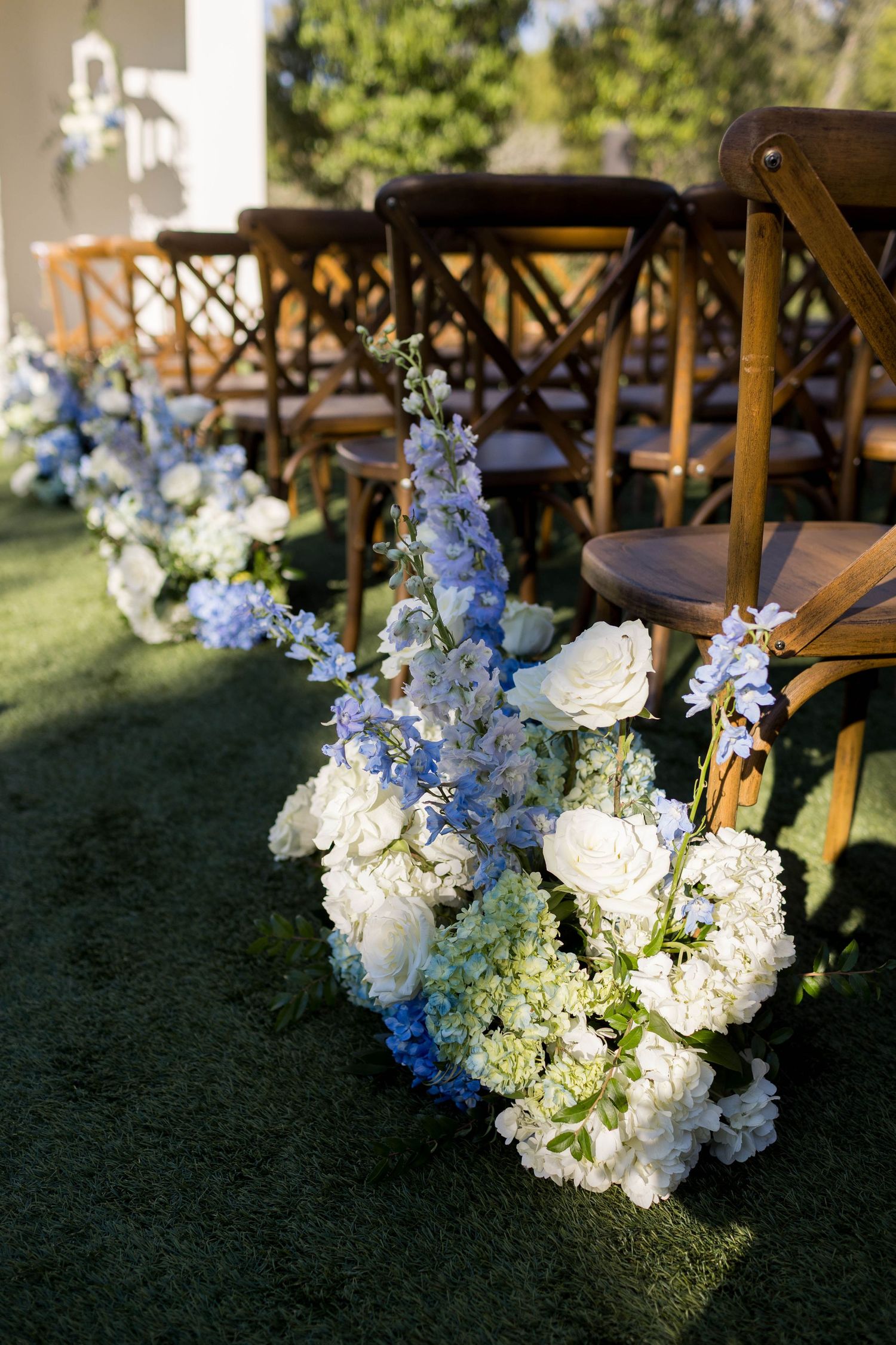 Wedding aisle lined with white hydrangea and blue delphinium floral arrangements beside rustic wooden cross-back chairs on green lawn.