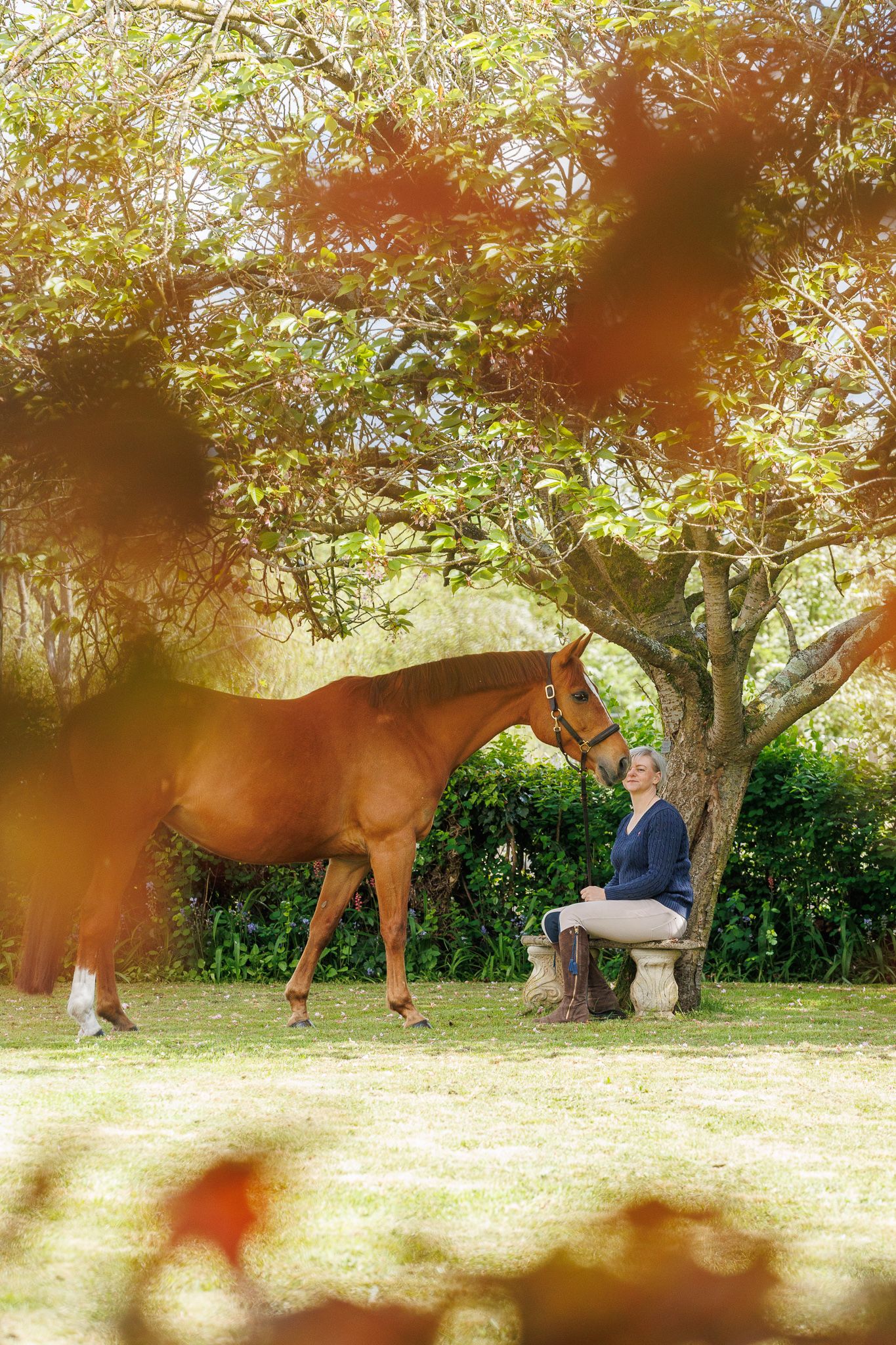 A brown horse and rider in a white sweater pose together against autumn foliage in soft natural lighting.
