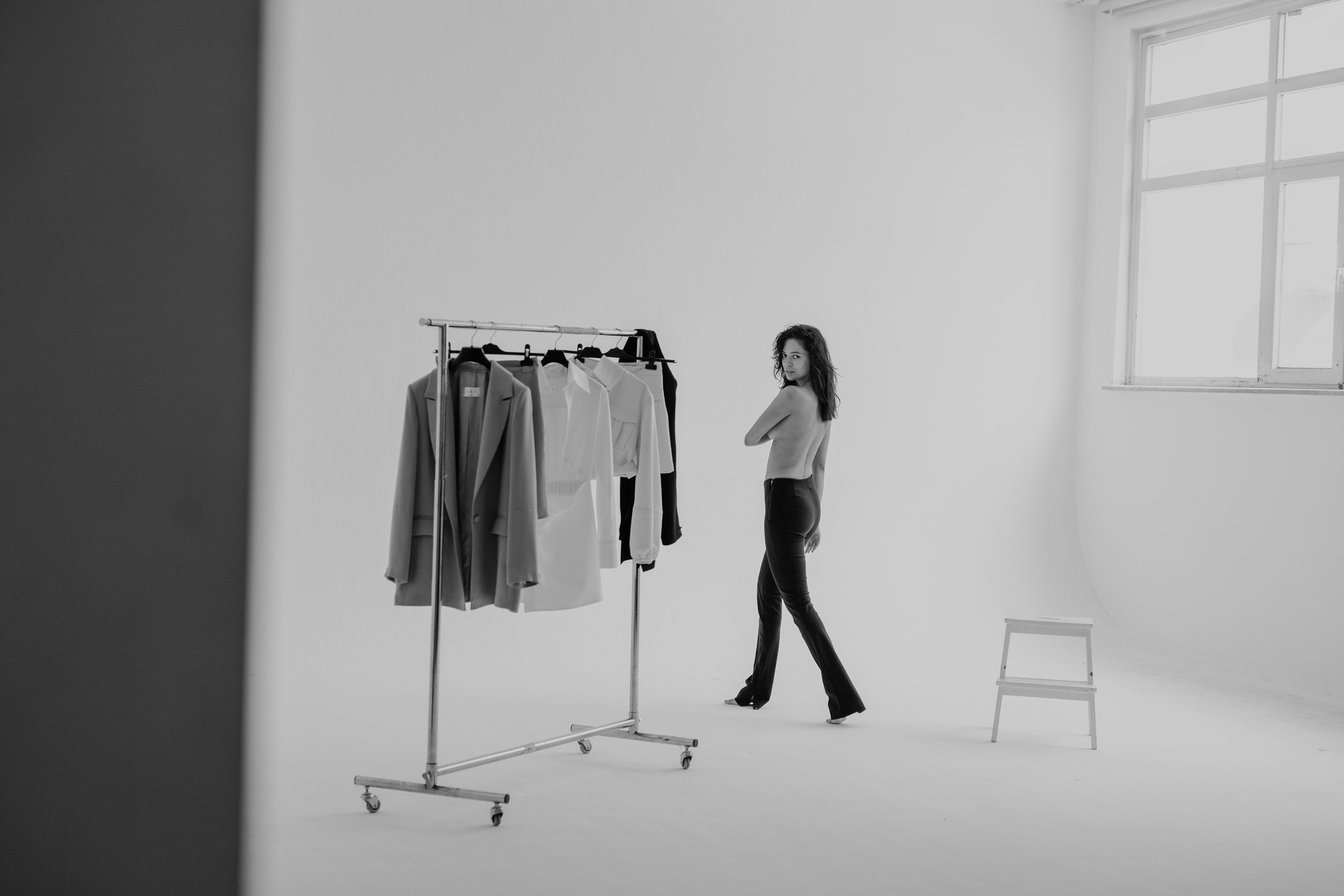 Woman in black pants stands near a clothing rack with garments in a minimalist white studio setting.